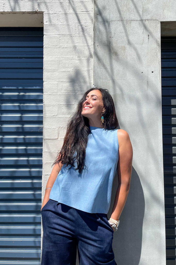 woman wearing aqua linen summer shell top from earn and learn with denim culottes leaning against concrete wall