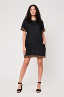 woman with curly hair looking at camera with one arm by her side and one hand in pocket wearing a black thigh length dress with leopard elastic trim, black heels against a white backdrop.