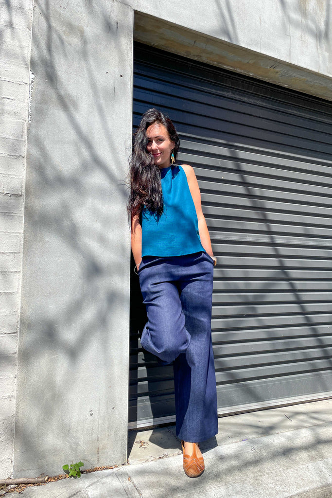 woman wearing peacock blue linen summer shell top from earn and learn with denim culottes leaning against concrete wall