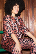 woman with curly hair wearing a black top and pants with copper curl print sitting with hands on knees against a wooden back drop