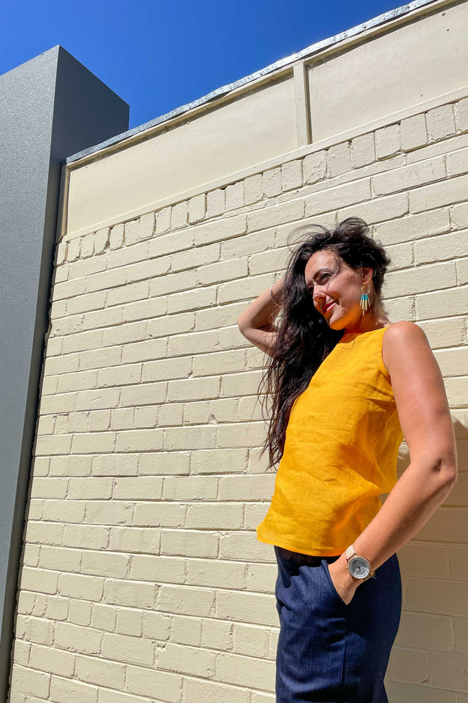 woman wearing sunshine yellow linen summer shell top from earn and learn with denim culottes leaning against concrete wall