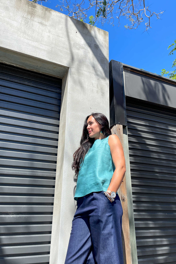 woman wearing turquoise linen summer shell top from earn and learn with denim culottes leaning against concrete wall