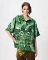 A model wearing a green short-sleeve button-up shirt with an abstract watercolor-style print in various shades of green, paired with olive green pants, against a plain light gray background.