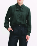 Person wearing a dark green blouse with a bow tie and dark pants on a white background