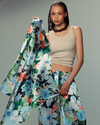 A model wearing a beige ribbed tank top and matching floral print wide-leg pants, holding up a matching floral print fabric panel against a solid gray studio background.