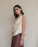 Woman wearing a cream sleeveless top with a draped design against a plain background.