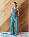 Woman wearing a grey tank top and teal pants standing against a wooden wall.