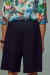 A person wearing black pleated shorts and a colourful floral print blouse, shown from the back against a plain light gray background.