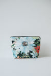A small zippered cosmetic pouch with a vibrant floral print featuring a large light blue flower, green leaves, and red accents, set against a plain light gray background.