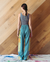 Woman wearing a grey tank top and teal pants standing against a wooden wall.