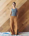 Woman wearing a grey tank top and brown pants against a wooden wall.