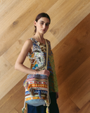 Model wearing a colourful sleeveless top and patterned bag against a wooden wall.