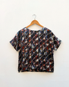 A short-sleeve top with a diagonal striped pattern in black, white, red, and blue, hanging on a wooden hanger against a white wall.
