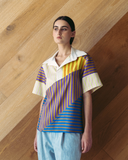 Model wearing a colourful geometric-patterned shirt against a wooden wall.