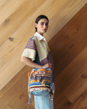 Model wearing a colourful top and carrying a patterned bag against a wooden wall.
