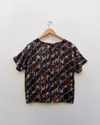 A short-sleeve top with an abstract vertical stripe pattern in black, orange, white, and red, hanging on a wooden hanger against a plain white wall.