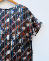 A short-sleeve top hanging on a wooden hanger against a white wall. The shirt features a complex abstract collage print with vertical black and white stripes overlaid on a background of orange, red, blue, and white abstract shapes.