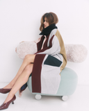 Woman sitting on a modern chair wearing a long, patterned coat against a white background