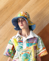 Person wearing a colourful patterned shirt and bucket hat against a wooden wall.