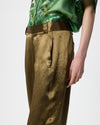 A close-up of a model wearing textured metallic olive-gold wide-leg trousers, paired with a green patterned top, against a plain light gray background.
