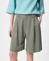A person wearing pleated olive green shorts and a light blue blouse, shown from the chest down against a plain light background.