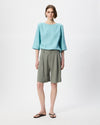 A model stands against a light gray studio background wearing a light blue three-quarter sleeve top, olive green pleated shorts, and black flip-flop sandals.