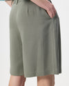 Close-up of a person wearing olive green tailored shorts with a side pocket and pleated front, shown against a light gray studio background.