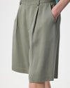Close-up of a person wearing pleated knee-length shorts in a muted sage green color, with a hand in the side pocket, against a plain light background.