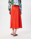A person shown from the back wearing a bright orange-red midi skirt, a floral long-sleeve top, and black sandals against a plain light gray studio background.