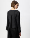 A woman shown from the back wearing a black long-sleeve top with a wide boat neckline and black textured trousers, photographed against a plain light gray studio background.