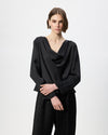 A model wearing a black draped long-sleeve top and black textured trousers, standing against a plain light gray studio background.