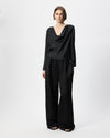 A model stands against a light gray studio background wearing a black draped long-sleeve top and matching black wide-leg trousers with a subtle textured pattern.
