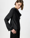A model wearing a black long-sleeve blouse with a draped, layered collar and a matching black quilted skirt, photographed against a clean white studio background.