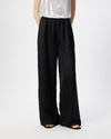 A model wearing black wide-leg trousers with a subtle textured diamond pattern, paired with a shiny white top and black sandals, shown from the waist down on a white studio background.