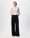 A model stands against a light gray studio background wearing a white sleeveless top and black wide-leg trousers.