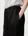 Close-up of a person wearing black wide-leg trousers with a subtle diamond grid pattern, paired with a white top, against a light gray background.