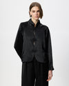 A model wearing a black satin-like cropped jacket with frog closures and black trousers, photographed against a plain light grey studio background.