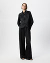 A model stands against a light gray studio background wearing a black textured two-piece outfit consisting of a long-sleeve cropped jacket with a mandarin collar and wide-leg trousers.
