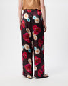 A model wearing wide-leg trousers with a dark burgundy base and a vibrant multicolor floral print in red, pink, blue, and cream, paired with a beige top and black sandals against a white studio background.