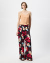 A model wearing wide-leg trousers with a dark burgundy base and a vibrant multicolor floral print in red, pink, blue, and cream, paired with a beige top and black sandals against a white studio background.