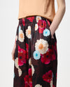 A model wearing wide-leg trousers with a dark burgundy base and a vibrant multicolor floral print in red, pink, blue, and cream, paired with a beige top and black sandals against a white studio background.