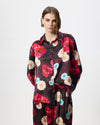 A model stands against a light grey studio background wearing a burgundy silk satin long sleeve shirt with a vibrant red, pink, and light blue floral print. 