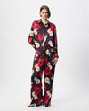 A model stands against a light grey studio background wearing a burgundy silk satin long sleeve shirt and wide leg pants with a vibrant red, pink, and light blue floral print. 