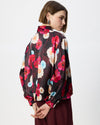 A woman shown from the back wearing a sheer black satin blouse with a vibrant red, pink, and light blue floral print, paired with burgundy trousers, against a plain white studio background.