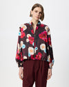 A model wearing a long-sleeve button-up blouse with a vibrant multicolor floral print on a dark background, paired with burgundy trousers, shot against a plain light gray studio background.