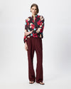 A model stands against a light gray studio background wearing a long-sleeve button-up blouse with a dark floral print and deep burgundy wide-leg trousers.