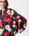 A close-up of a model wearing a satin bomber jacket with a vibrant floral print in red, pink, white, and light blue over a dark base.