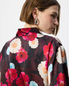 A close-up side profile of a model wearing a dark floral silk blouse with vibrant pink, red, and white flowers, paired with a silver hoop earring.