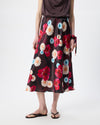 A model is shown from the waist down wearing a dark burgundy midi skirt with a vibrant red, pink, and light blue floral print. The model is also wearing a dark brown top and black flip-flop sandals against a plain white studio background.