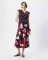 A model stands against a light gray studio background wearing a sleeveless dark brown draped top and a dark midi skirt with a large red, pink, and white floral print. She is wearing black flip-flop sandals.